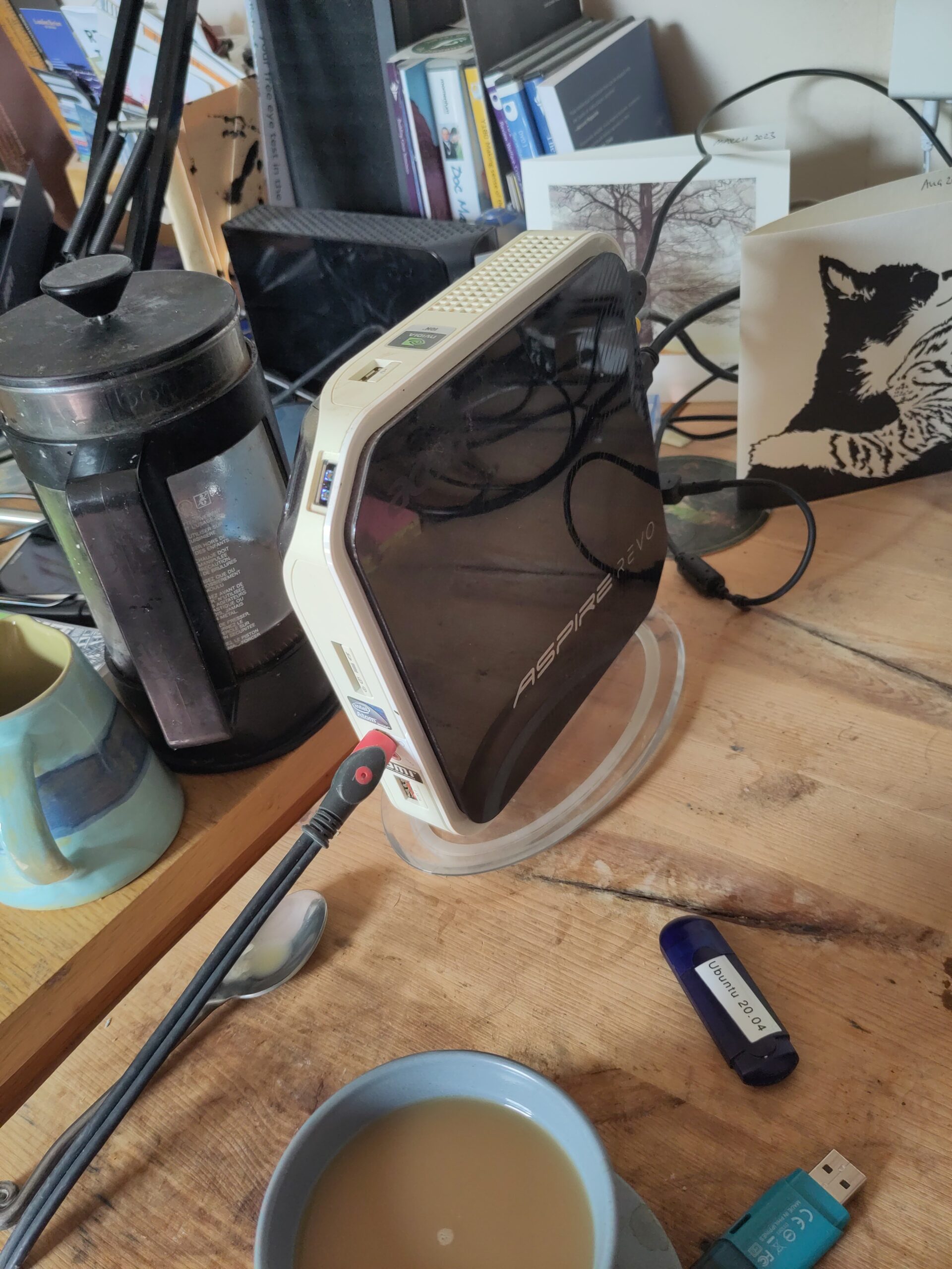 A small computer of peculiar design sits on a chaotic desk, with a pot of coffee, a cup and saucer, a jug of milk, and a memory stick labelled, "Ubuntu 20:04"
