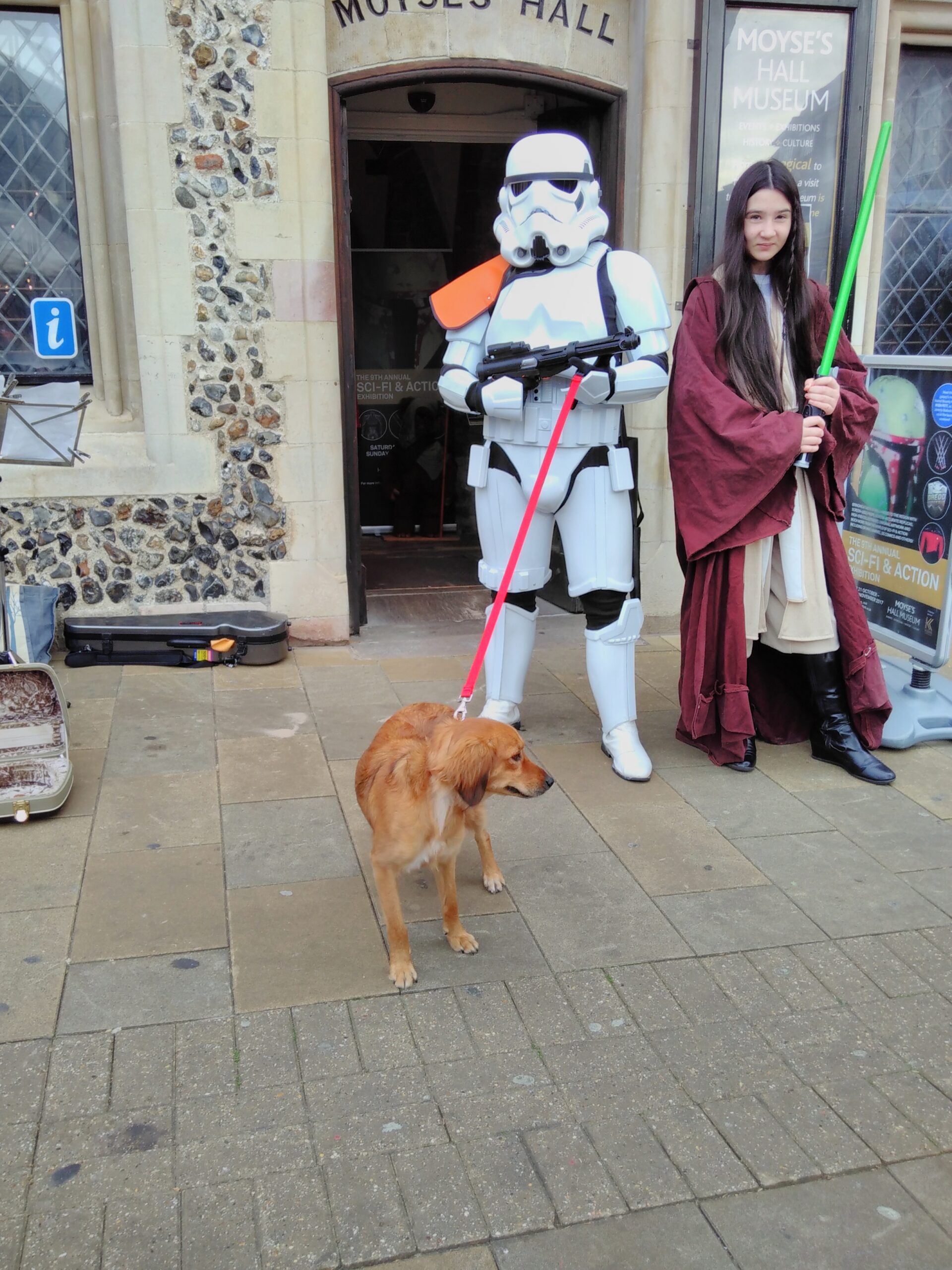 An Imperial Stormtrooper holds the lead of a dog and a teenage girl dressed as a jedi wields a plastic light sabre in front of a mediaeval flint-walled building. Yes, really.