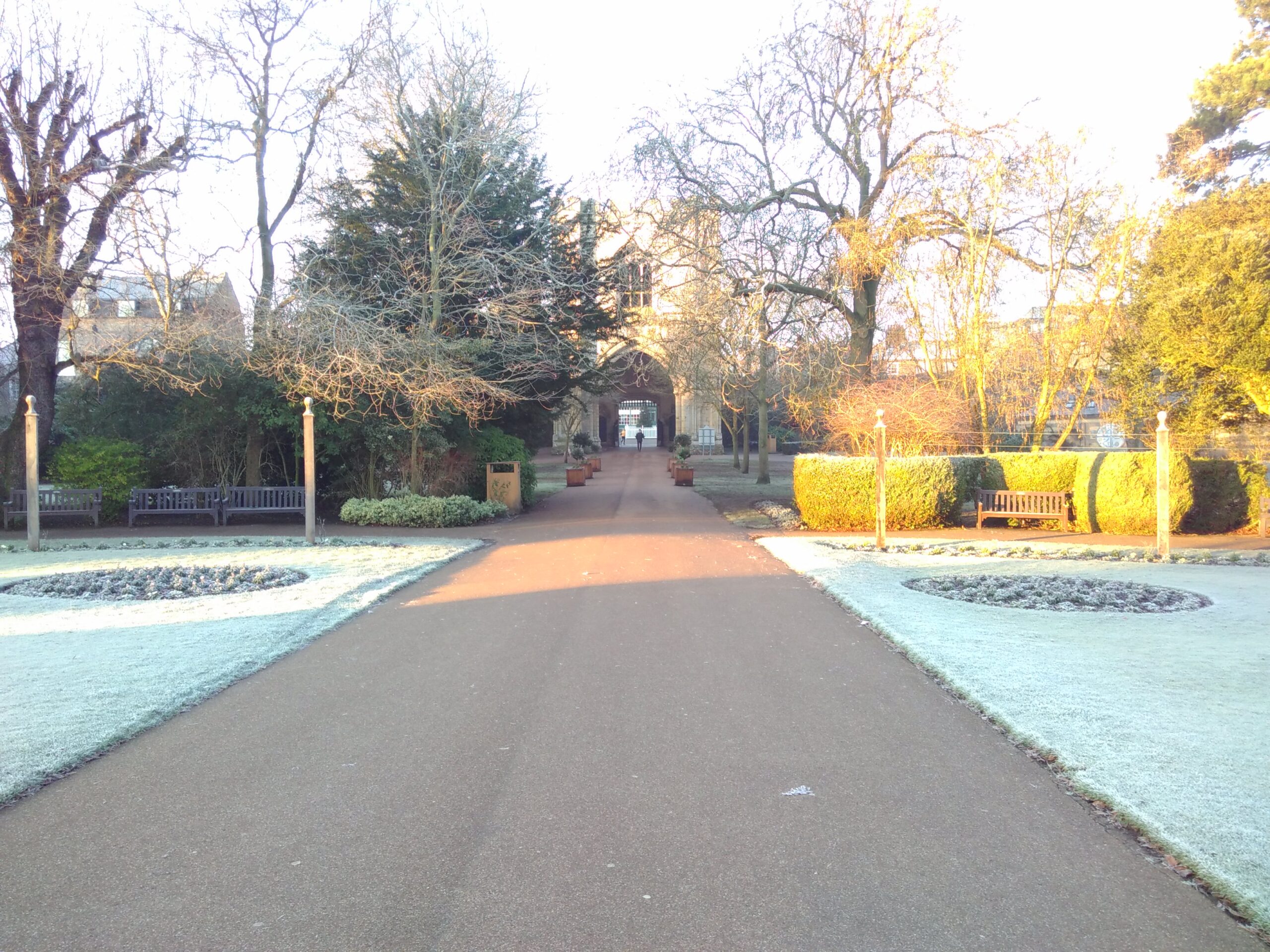 A path between frost-covered park lawns towards a mediavel stone tower beyond hedges and trees.