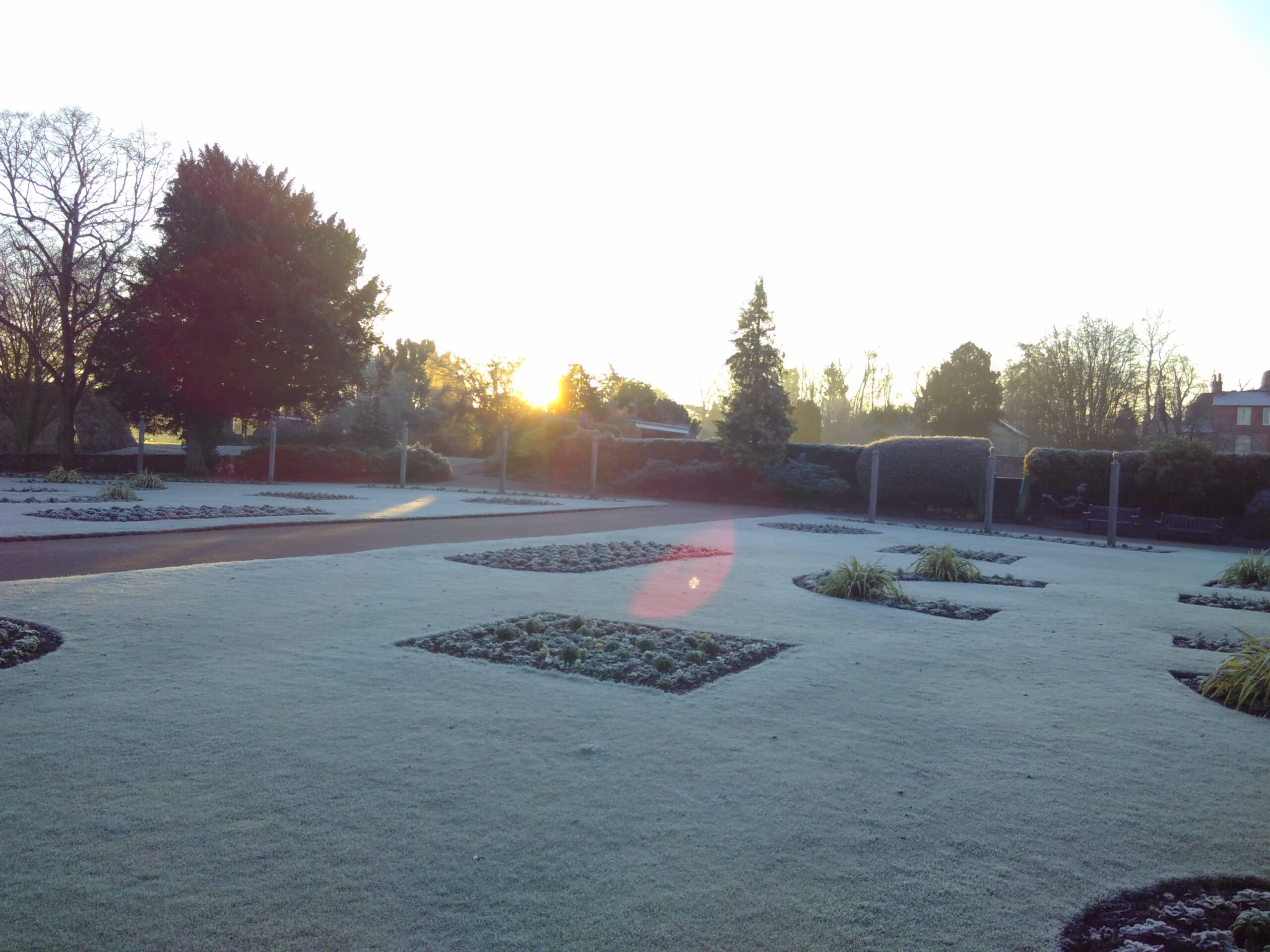 The sun rises beyond hedges on a frosty morning in a park.