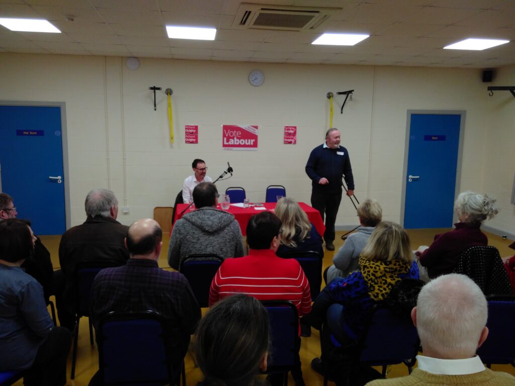 A meeting in a community centre. An audience faces a stage with two men at a Labour Party branded table.