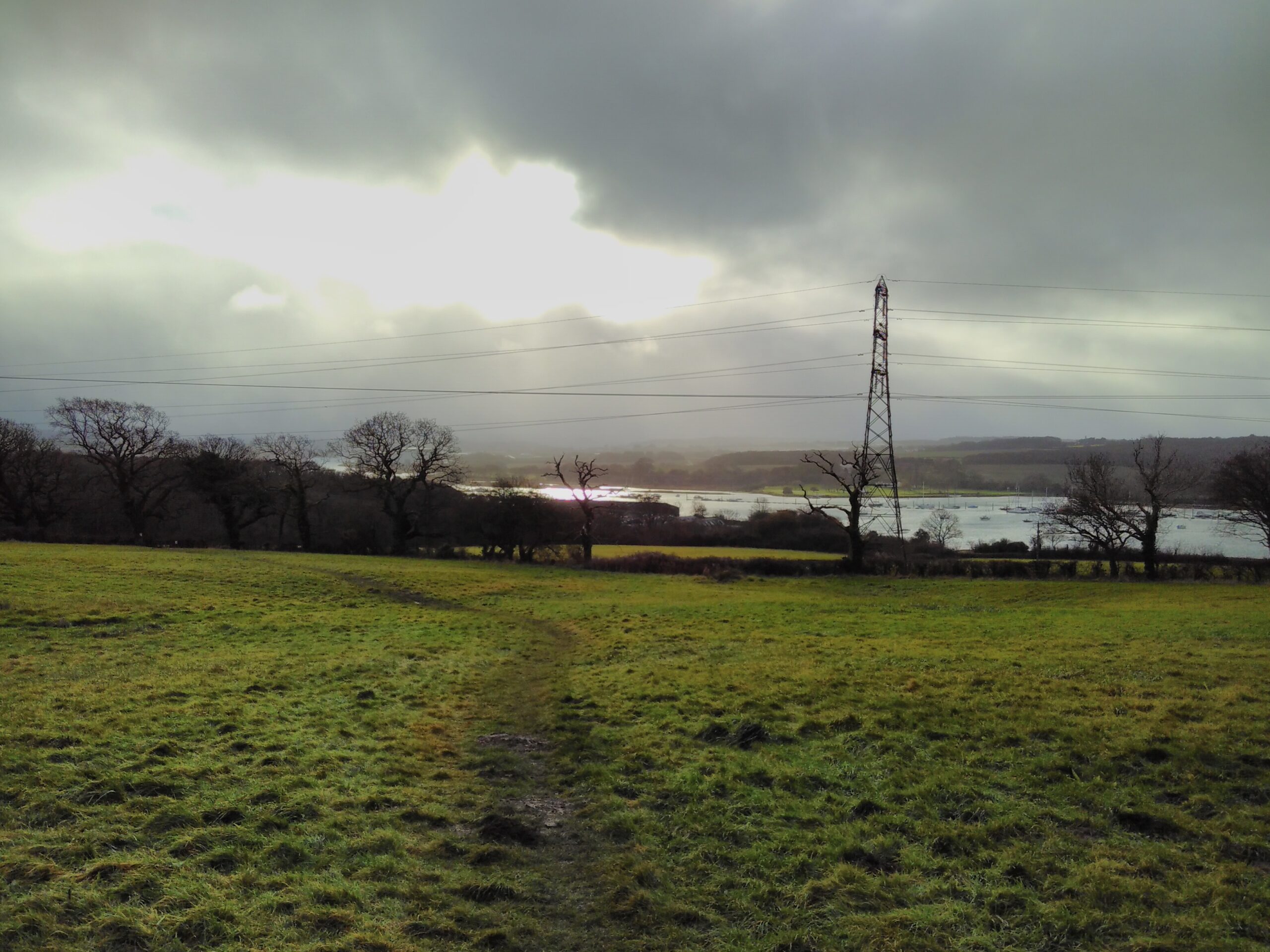 A muddy path crosses a field sloping down to a broad river, with an electrical pylon next to a wintry woodland in the distance. The sky is grey clouded with a yellow lemon-sun breaking through.