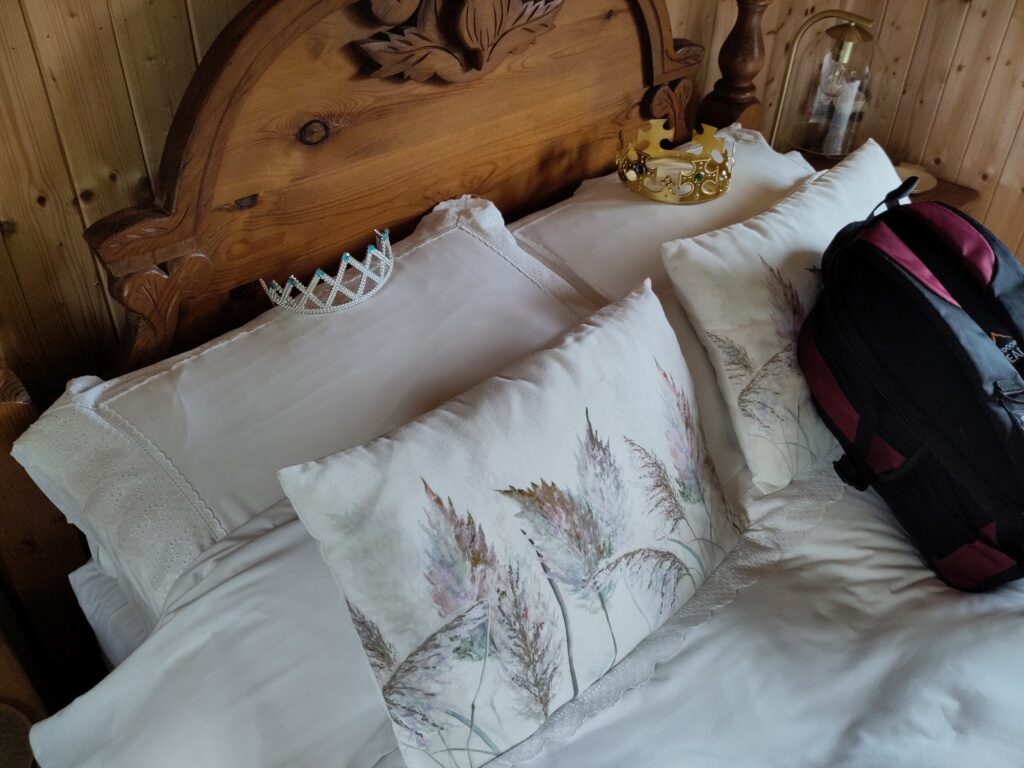 Pillows on a freshly made bed, with plastic crown and tiara. The bedhead is carved wood and the walls are pine clad.