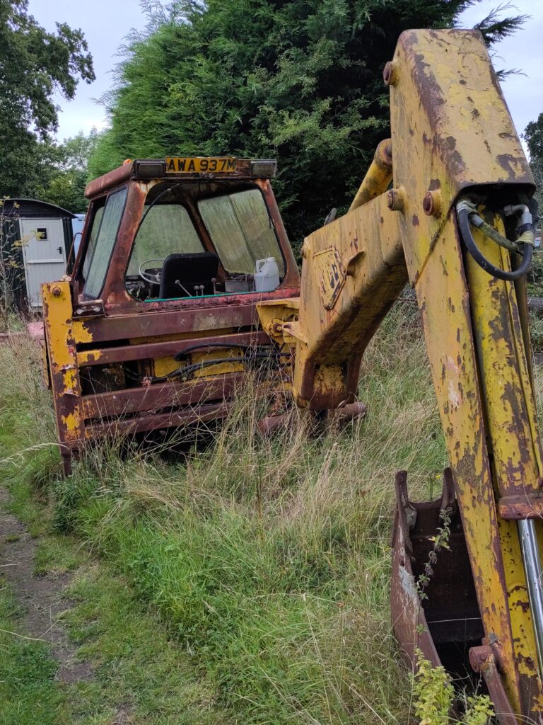 A rusting JCB digger, photographed from behind, its bucket arm stretched towards the camera like a dinosaur's neck.