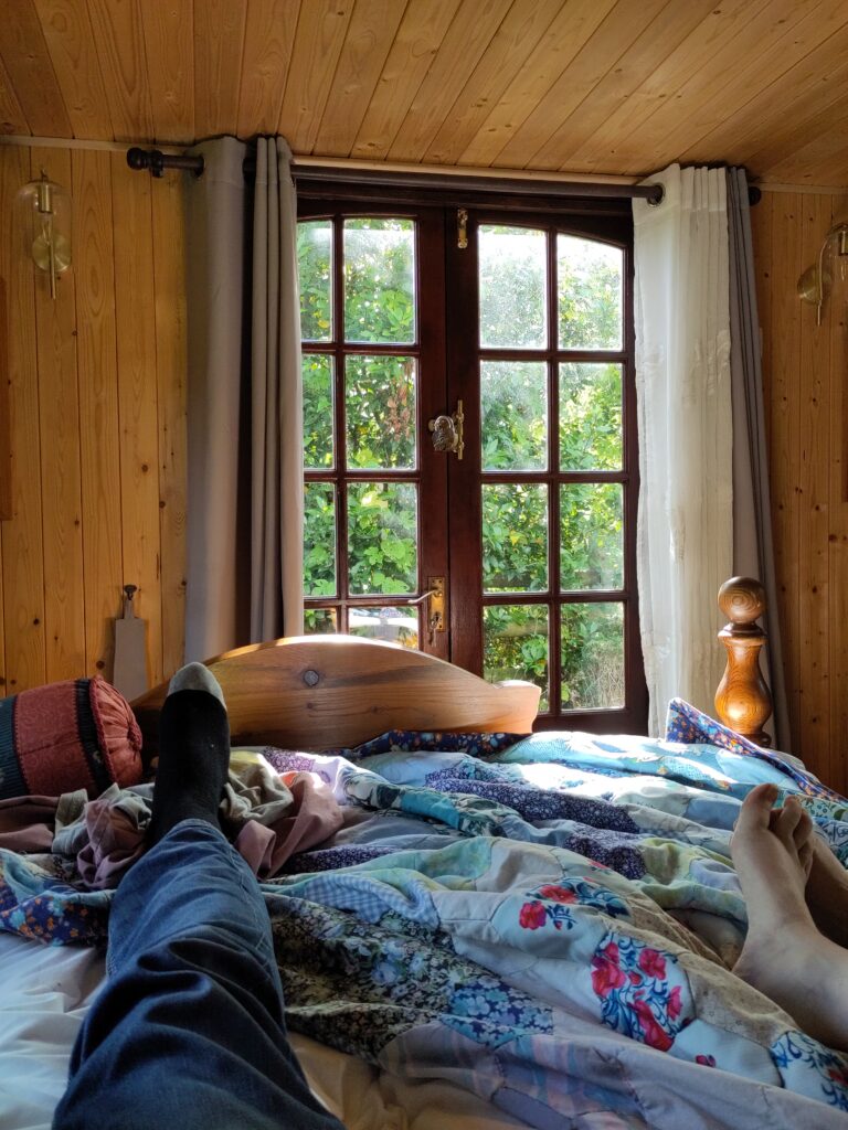 A couple's legs are stretched out on a bedspread. He wears jeans and socks; her feet are bare. At the end of the bed are a pair of French windows looking out at eh yard surrounded by greenery. Sun glows against the windows.