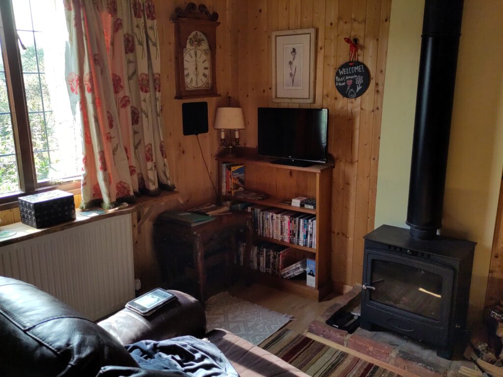 The cabin sitting room in the morning. An e-bbok reader sits on the arm of a leather sofa, the stove is dark, the telly is off. A small circular chlakboard, hung on the wall with a ribbon, reads, "Welcome! Peter, Amanda & Hazel."