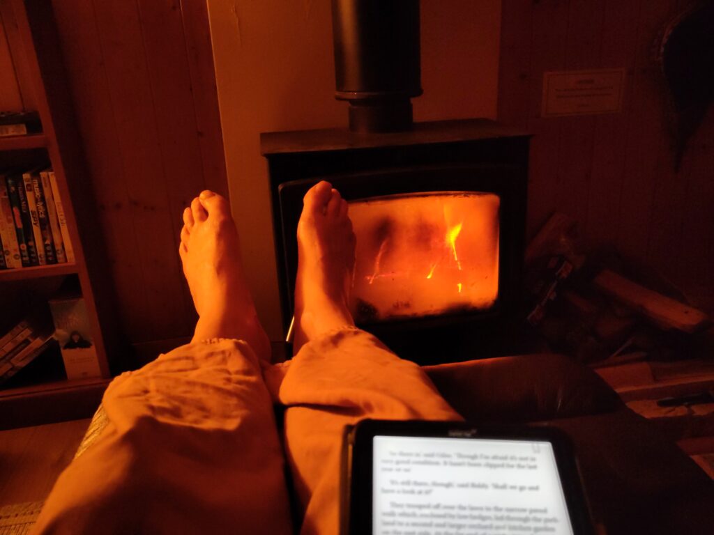 A man's legs and bare feet are framed by the light of a wood burning stove. On his lap is an e-book. Text is visible but indecipherable.