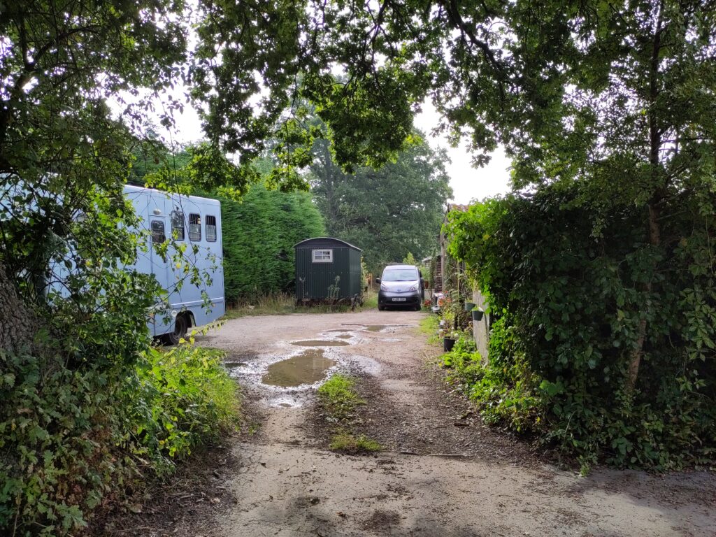 A rough, puddled lane is bordered by trees and hedges. At the end are a wheeled shepherd's hut and a small modern van. A horse box truck is parked to the side.